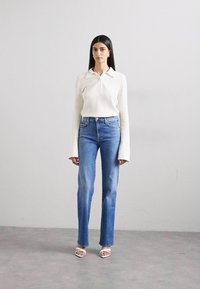 White ribbed long-sleeve shirt with a collar, paired with light blue straight-leg jeans and white open-toe heels against a neutral background.
