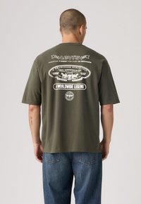 Λαδί βαμβακερό t-shirt με λευκό γραφικό τύπωμα στο πίσω μέρος, διαθέτει λεπτομέρειες και λογότυπα μάρκας, άνετη εφαρμογή, κοντά μανίκια.