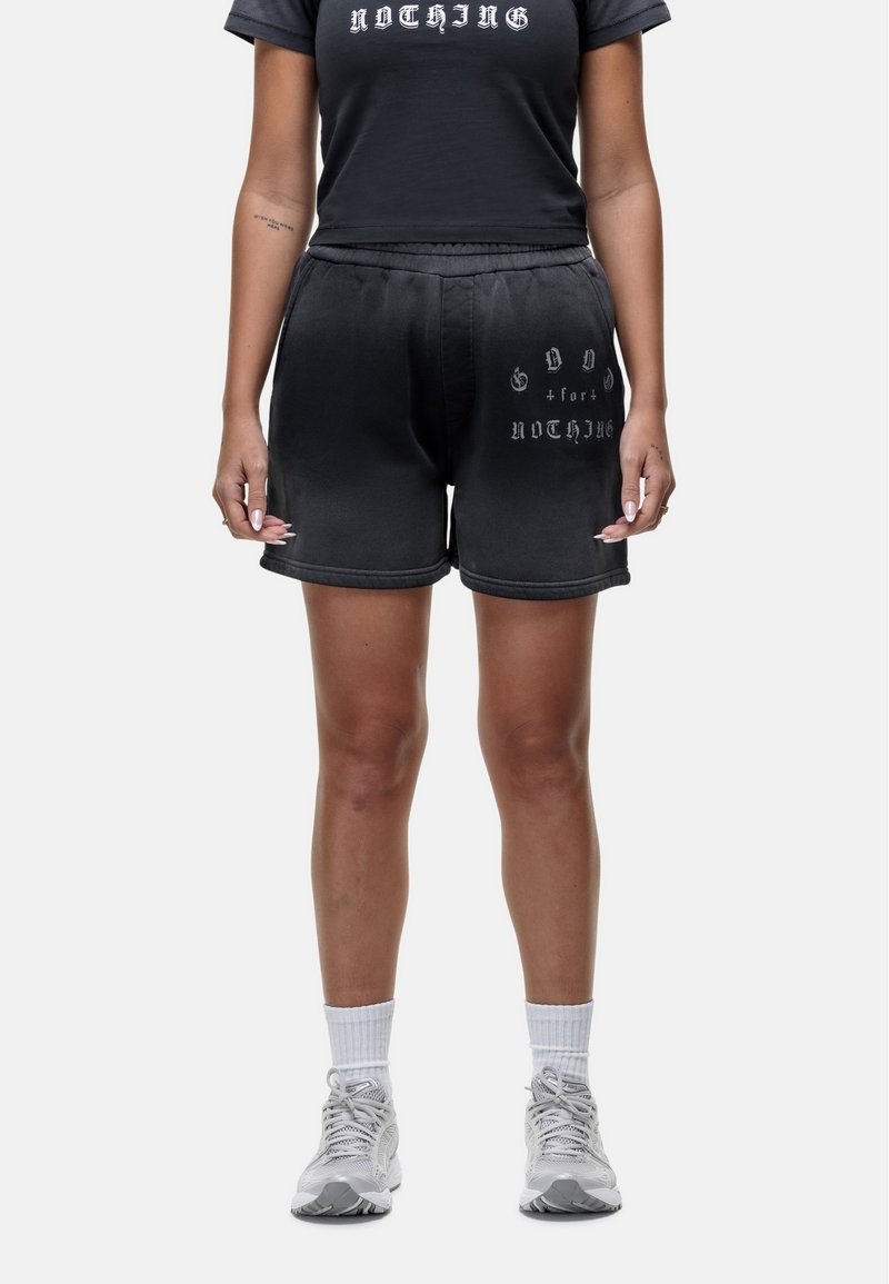Shorts en coton noir avec une taille élastique, ornés d'imprimés en texte blanc et d'une coupe décontractée. Associés à des chaussures de sport blanches et des chaussettes.