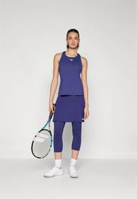 Γυναίκα με μπλε αμάνικο τοπ, φούστα πάνω από κολάν μέχρι το γόνατο, κρατώντας ρακέτα τένις, φορώντας λευκά παπούτσια και κάλτσες, στέκεται μπροστά σε απλή τοιχογραφία.