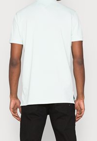 Polo shirt court à manches courtes vert menthe clair avec un col en V, texture lisse, fentes latérales et un détail de marque subtil à l'ourlet. Porté avec un pantalon noir.