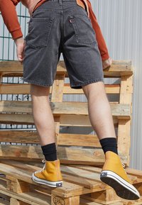 Shorts de mezclilla gris con un aspecto desgastado, que cuentan con bolsillos traseros. Una persona lleva zapatillas altas amarillas y calcetines negros, de pie sobre palets de madera.