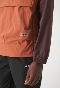 Oransje og bordeaux genserjakke, myk stoff, elastiske mansjetter, logo-patch på siden, har en brystlomme med klaff.