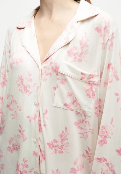 Femme portant un haut de pyjama blanc boutonné avec des motifs floraux rose clair et une poche poitrine, visible du cou jusqu'au milieu du torse.