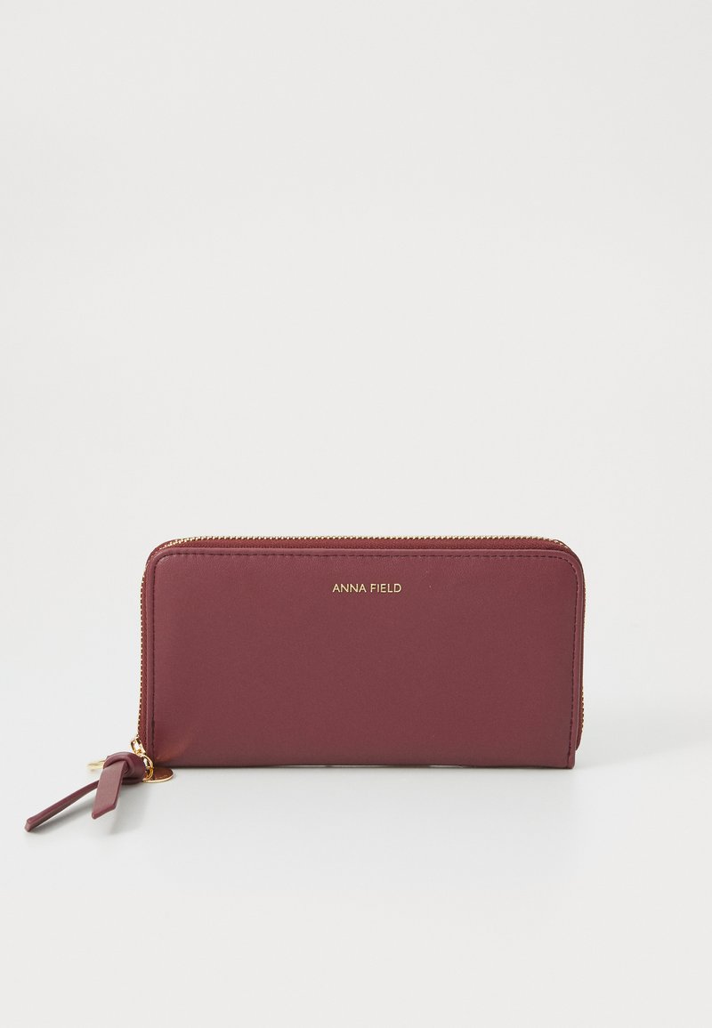 Carteira burgundy com fecho em zíper, de textura suave, com hardware dourado e logótipo em relevo "ANNA FIELD" na frente. Apresenta um pequeno pompom no zíper.