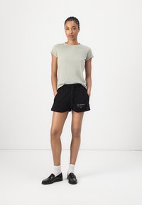 T-shirt verde chiaro con maniche arrotolate, abbinata a pantaloni corti neri con cordoncino e tasche laterali. Scarpe nere e calzini bianchi.