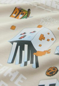 Bela pikselizirana pajek z rdečimi očmi in črnimi nogami, obdana z oranžnimi bloki in pikseliziranimi predmeti na bež tkanini.