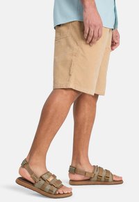 Μπεζ σορτς από κορδόνι με πλαϊνή τσέπη, συνδυασμένα με καφέ σανδάλια με λουράκια και ρυθμιζόμενες αγκράφες και υφασμένα πέλματα.