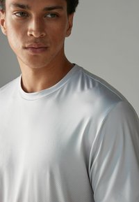 Lys grå atletisk t-shirt med en glat tekstur, rund halsudskæring og subtil honningcomb-mønster. Stoffet synes at være fugttransporterende og tætsiddende.