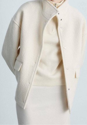 Veste en mélange de laine crème avec col montant, fermeture à pression et poches latérales, portée sur un haut en tricot côtelé assorti et une jupe ajustée.