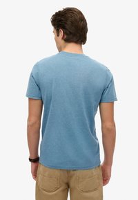 Ung mand med kort brunt hår iført en blå kortærmet t-shirt og beige bukser, set bagfra mod en hvid baggrund.