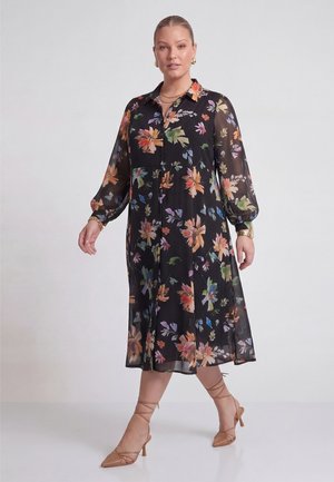 Kvinde iført en sort midikjole med farverigt blomsterprint, lange gennemsigtige ærmer, knapper foran og brune snørestilethæle, stående på en hvid gulv.