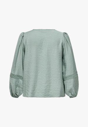 Blusa texturizada verde claro con mangas largas y voluminosas. Presenta un sencillo escote redondo y detalles trenzados en los puños de las mangas.
