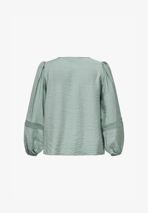 Világos zöld, texturált blúz hosszú, voluminózus ujjal. Egyszerű, kerek nyakkivágással rendelkezik, és fonott díszítésekkel a ujjgömbökön.