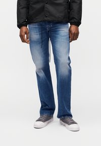 Άνδρας με ξεθωριασμένο μπλε τζιν, γκρι πάνινα αθλητικά παπούτσια και μαύρο μπουφάν, στέκεται μπροστά σε απλό λ ευκό φόντο.