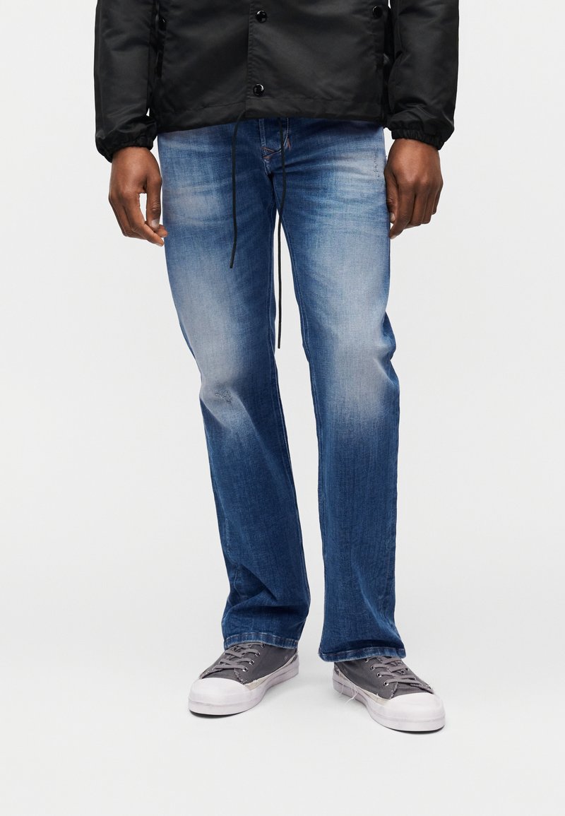 Άνδρας με ξεθωριασμένο μπλε τζιν, γκρι πάνινα αθλητικά παπούτσια και μαύρο μπουφάν, στέκεται μπροστά σε απλό λευκό φόντο.