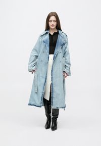 Mulher em pé a usar um casaco longo de ganga azul claro por cima de um top preto, saia branca com botões e botas pretas até ao joelho, sobre um fundo branco.