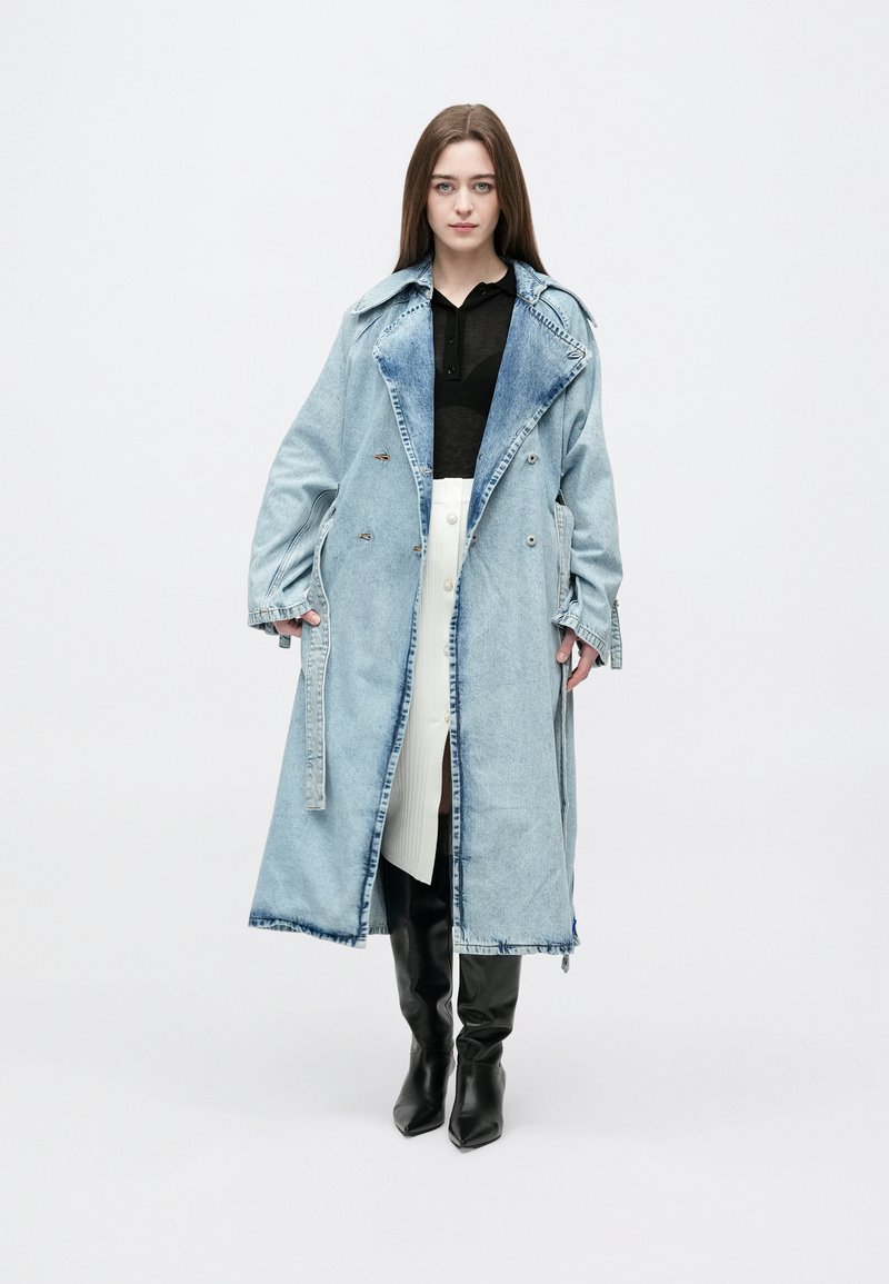 Mulher em pé a usar um casaco longo de ganga azul claro por cima de um top preto, saia branca com botões e botas pretas até ao joelho, sobre um fundo branco.