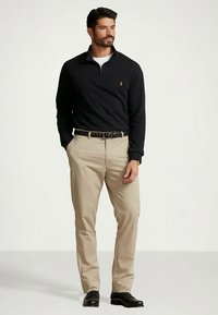 Moški stoji oblečen v črno pulover z četrtim zadrgo, bež hlače, črn pleten pas in črne čevlje, roke v žepih, gleda desno.