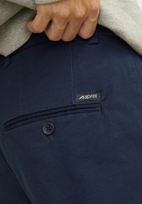 Hånd justerer lomme på marineblå bukser med sort knap og Aprel-mærke, iført grå strikket sweater.