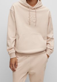 Sudadera beige con bolsillo canguro en la parte frontal, capucha con cordones y logo en relieve. Presenta puños y dobladillo de canalé. Textura de tela suave y lisa.