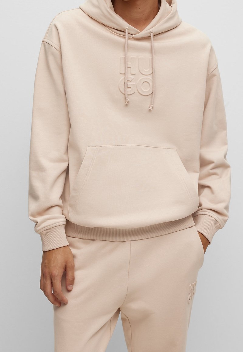 Sudadera beige con bolsillo canguro en la parte frontal, capucha con cordones y logo en relieve. Presenta puños y dobladillo de canalé. Textura de tela suave y lisa.