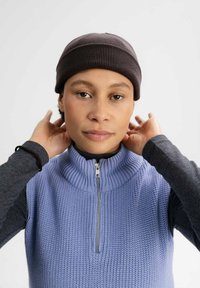 Gehaakte grijze beanie, geribbelde structuur; lichtpaarse mouwloze trui met hoge kraag en ritsdetails. Donkere lange mouwen top eronder.