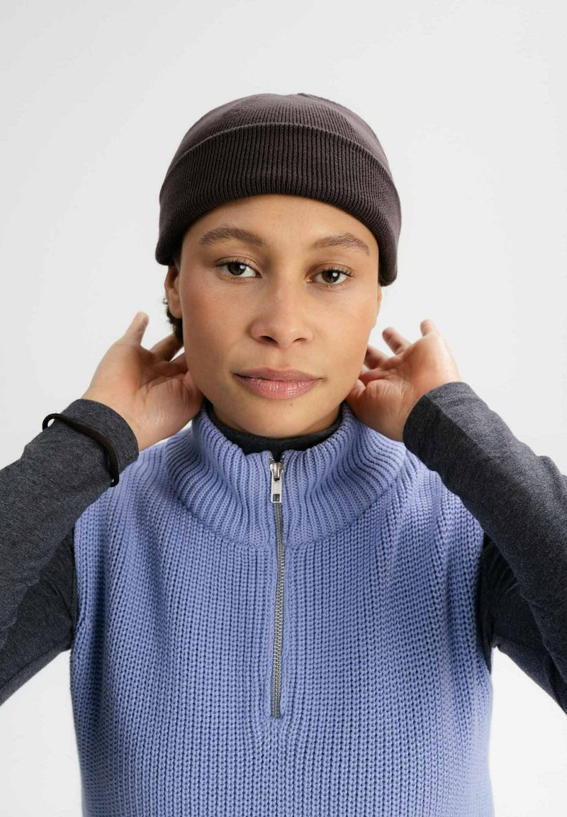 Gehaakte grijze beanie, geribbelde structuur; lichtpaarse mouwloze trui met hoge kraag en ritsdetails. Donkere lange mouwen top eronder.