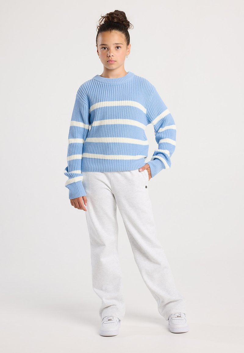 Girl with curly hair in a bun wearing a blue and white striped sweater, white sweatpants, and white sneakers standing with one hand in pocket.