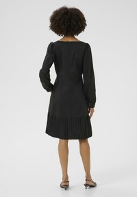 Robe noire avec une jupe plissée, des manches longues et un col rond. Présente des accents de boutons sur les poignets et une fermeture éclair invisible au dos.
