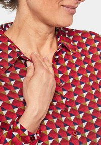 Camicetta rossa con un motivo geometrico di triangoli in verde, bianco e blu navy. Presenta un colletto e maniche lunghe con chiusure a bottone.