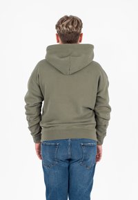 Felpa verde oliva con un ampio cappuccio, cuciture visibili e polsini e orli a costine. Indossata con jeans in denim blu.