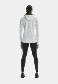 Weißer sportlicher Hoodie mit Kapuze, langen Ärmeln und reflektierenden Akzenten; kombiniert mit schwarzen Leggings und robustem Schuhwerk.