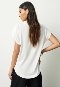 Bílé tričko s krátkými rukávy s volným střihem, zaobleným lemem a ohrnutými rukávy, v kombinaci s černými kalhotami. Hladká textura a minimalistický design.