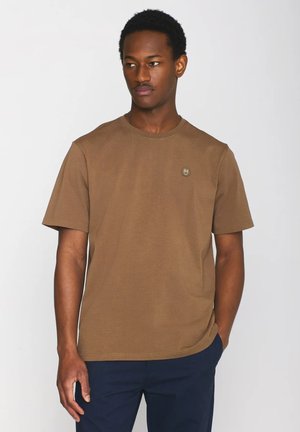 Uomo con camicia marrone a maniche corte con piccolo emblema sul petto e pantaloni scuri, in piedi contro uno sfondo chiaro uniforme, guarda verso sinistra.