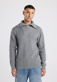 Hombre con cabello corto y oscuro, vistiendo un suéter gris de canalé con cremallera y jeans azul claro, de pie contra un fondo blanco liso.