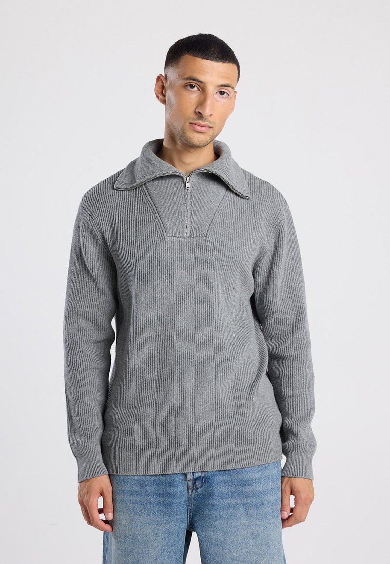 Hombre con cabello corto y oscuro, vistiendo un suéter gris de canalé con cremallera y jeans azul claro, de pie contra un fondo blanco liso.