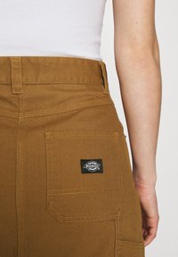 Gros plan sur une personne portant un pantalon marron Dickies avec une poche arrière et une chemise blanche, mettant l'accent sur la poche et le logo.