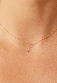 Collier en or avec un petit pendentif en forme de lettre "j" minuscule. La chaîne est fine et délicate, reposant sur une peau lisse.