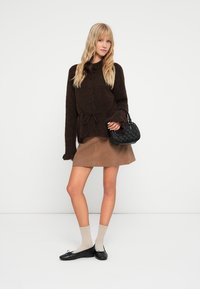 Cárdigan marrón texturizado con cierre de botones y cinturón en la cintura, combinado con una falda beige. Bolso negro y calcetines claros con zapatos negros.