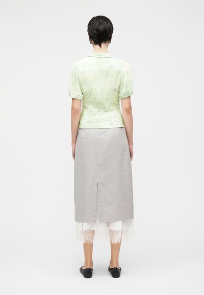 Ανοιχτό πράσινο μπλουζάκι με φουσκωτά μανίκια και εφαρμοστή μέση, σε συνδυασμό με μακριά γκρι φούστα με σκίσιμο. Λευκές δαντέλες φαίνονται στο κάτω μέρος.
