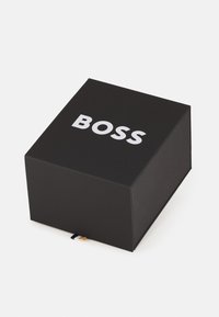 Čierna lepenková krabica s plochým viečkom, na vrchu s bielym textom "BOSS" a malou farebnou stužkou na boku.