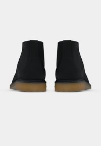 Ζευγάρι μαύρα μποτάκια αστραγάλου από σουέντ με τακούνια από καφέ κρεπ, φωτογραφημένα από πίσω πάνω σε λευκό απλό φόντο.