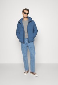 Doudoune bleue avec capuche, fermeture éclair à l'avant, portée sur un pull gris, associée à un jean bleu clair et des baskets marron. Lunettes de soleil incluses.