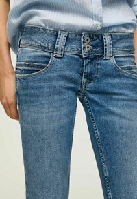 Jean en denim bleu avec une coupe slim, présentant deux poches avant, une fermeture à boutons, et des coutures contrastées. Le tissu semble légèrement usé.