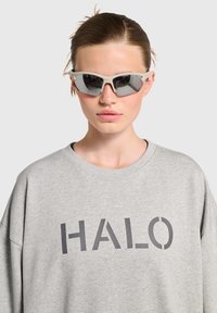 Solglasögon med en ljusgrå ram och spegelglas, bärs med en grå oversize sweatshirt med texten "HALO" i mörkgrått.
