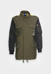 Olive green jacket with black sleeves, two chest pockets, and decorative accents. Features drawstring hem and patterned fabric details.