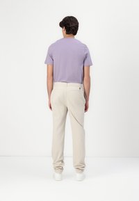T-shirt court à manches courtes de couleur lavande clair associé à un pantalon beige. La tenue est complétée par des baskets blanches et présente des coutures discrètes ainsi qu'un détail de logo.