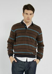 Brązowy sweter w paski z jasnoniebieskimi i granatowymi paskami, z granatowym kołnierzykiem i zapinanym dekoltem, noszony z białą koszulą pod spodem.