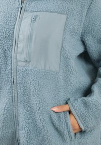 Giacca azzurro chiaro sfocata con una texture morbida, dotata di una tasca sul petto con zip e tasche laterali. Realizzata in materiale felpato, progettata per il calore.